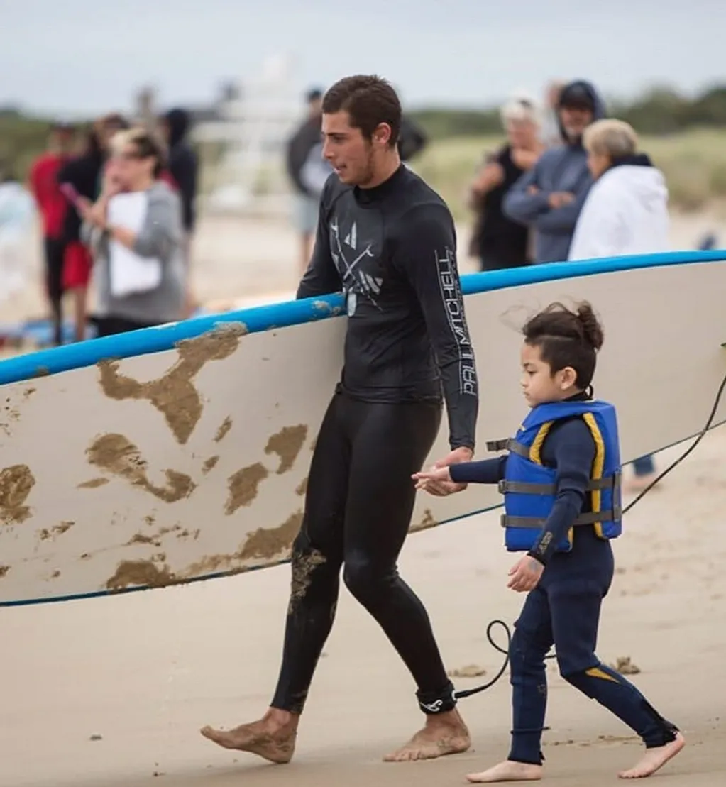 Ash, Strictly Surf instructor, walking the beach with a young student and surfboard in Amagansett